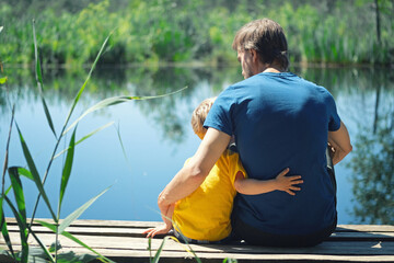 Happy father and son hugging sitting on wooden pier near lake on sunny summer day. Dad and child...