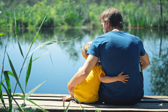 Happy father and son hugging sitting on wooden pier near lake on sunny summer day. Dad and child boy spend time together on wharf. Father's day concept.