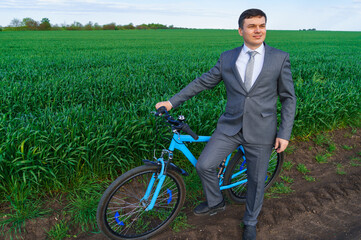 businessman with a bicycle poses in a wheaten field, green grass and blue sky as background