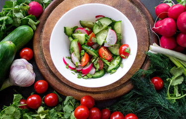 A pile of fresh, ripe vegetables: radishes, tomatoes, cucumbers, herbs, and garlic are arranged around a round wooden chopping board. White bowl vegetable salad, dressing with olive oil