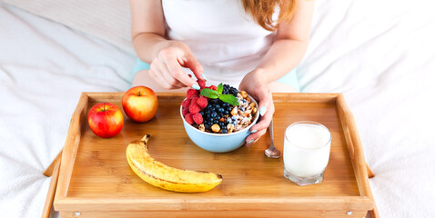 A young woman eats fresh raspberries, blueberry and granola for breakfast while sitting on bed. Healthy breakfast concept.