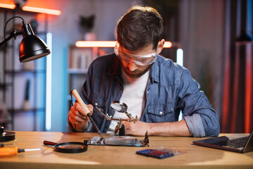 Focused bearded man in protective glasses sitting at table and soldering video card from modern...