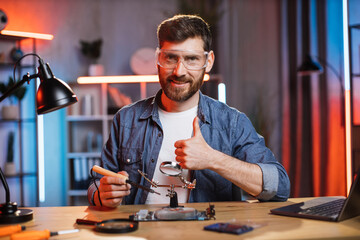 Caucasian young man in protective glasses showing thumb up, using soldering iron and magnifying glass while repairing display card from modern laptop. Concept of people and technology.