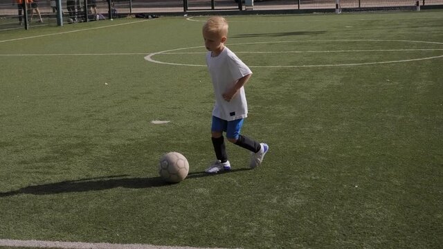 A little boy trains on the football field, practicing dribbling and feints.