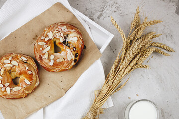 Caramel buns with raisins and almonds are lying on kraft paper in a tray. Next to it is a bouquet of wheat and rye ears, a mug with milk on a concrete background, selective focus.