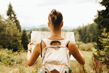 Hiking young woman traveler with backpack checks map to find directions in wilderness area, real explorer. Travel Concept - obrazy, fototapety, plakaty
