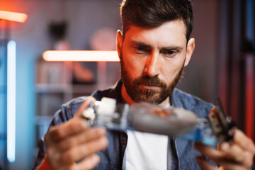 Close up of focused bearded man sitting at table and examining problem of non working display card from modern laptop. Young guy trying to fix details at home.
