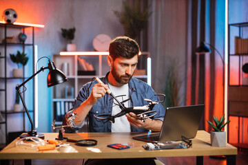 Fototapeta na wymiar Focused bearded man sitting at table and watching online tutorial about repairing of drone. Young guy looking on computer screen and soldering quadcopter.
