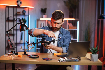 Plakat Concentrated bearded man using various tools for repairing modern quadcopter at home. Young caucasian guy in denim shirt fixing flying drone by himself.