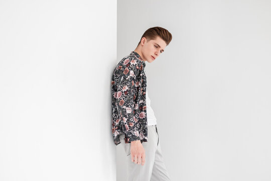 Elegant Pretty Young Man With Trendy Hairstyle In White T-shirt In Floral Vintage Shirt In Stylish Trousers Is Resting Near White Wall In Studio. Attractive Guy Model In Youth Fashion Clothes Indoors.