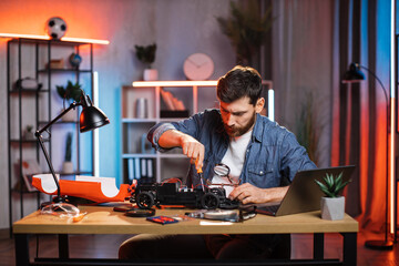 Bearded man using screwdriver and magnifying glass for fixing remote controlled toy car. Young guy sitting at table with various tools during repairing.