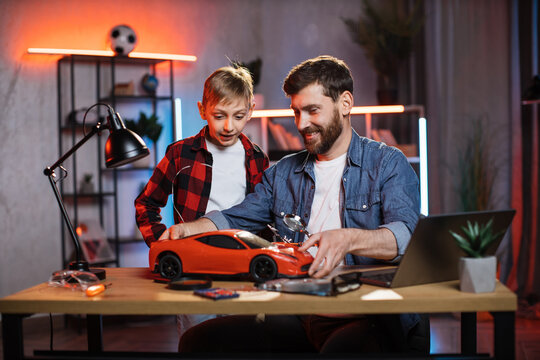 Happy young man with little boy successfully repaired remote controlled toy car together. Father with son in casual outfit spending time usefully at home.