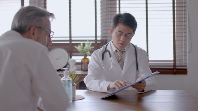 Senior Caucasian man consults with young Asian doctor. Doctor is describing health checking result to patient. medium close-up with slide movement.  