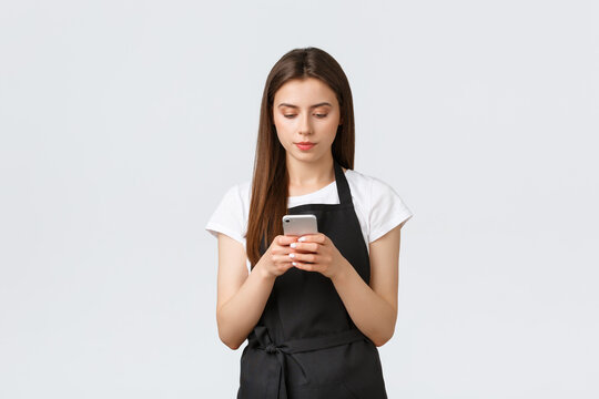 Grocery Store Employees, Small Business And Coffee Shops Concept. Serious Young Female Barista, Waitress On Break In Black Apron Using Smartphone, Looking Focused At Screen