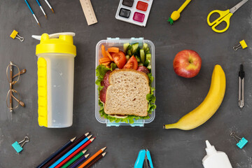 A school lunch box with a sandwich, vegetables, water and fruit on a gray concrete background. School supplies, stationery, and food. Flatly. From above.