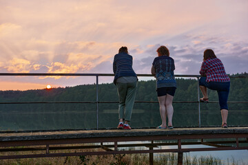 Trzy kobiety w zadumie obserwują malowniczy jesienny zachód słońca nad jeziorem - obrazy, fototapety, plakaty