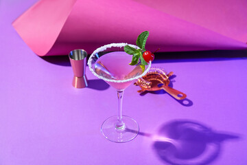 Glass of tasty cosmopolitan cocktail, shaker and strainer on color background