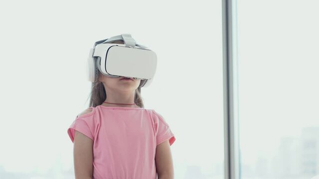 The girl is having fun in virtual reality glasses, stands against the background of the window and pretends that she is swimming. High quality 4k footage