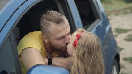 Handsome young bearded Caucasian man sitting in car sticking out of window as cute little girl coming kissing and hugging parent. Portrait of happy father enjoying travelling with daughter outdoors