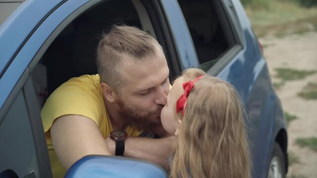 Handsome young bearded Caucasian man sitting in car sticking out of window as cute little girl coming kissing and hugging parent. Portrait of happy father enjoying travelling with daughter outdoors