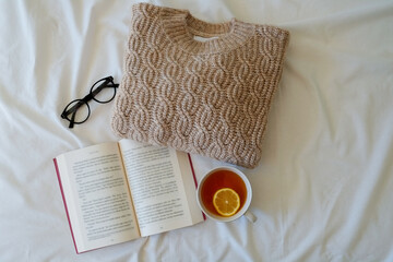 Knitted pastel color sweater perfectly folded on the bed. Open book, eyeglasses and tea with piece...