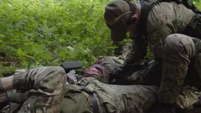 Combat medic taking first aid kit, providing emergency medical treatment to injured soldier while military group of equipped soldiers in camouflage covering companions in arms in forest area
