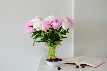 Close up shot of white kitchen counter with bouquet of beautiful peony flowers in glass vase, a book and a bowl of cherries. Reading and snacking. Copy space for text, close up, background.