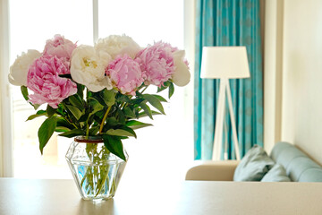 Close up shot of white counter with bouquet of beautiful white peony flowers in glass vase on foreground and blue textile couch on the background. Copy space for text, close up.