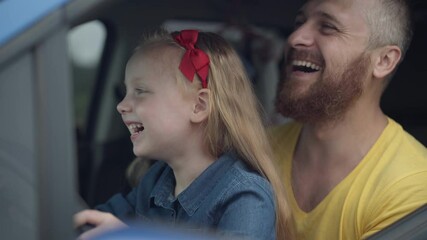 Close-up of excited happy little daughter sitting with father turning steering wheel in car laughing. Cheerful Caucasian cute girl and bearded handsome man enjoying leisure together in automobile