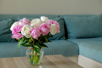 Close up shot of wooden coffee table with bouquet of beautiful white peony flowers in glass vase on foreground and blue textile couch on the background. Copy space for text, close up.