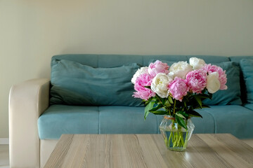 Close up shot of wooden coffee table with bouquet of beautiful white peony flowers in glass vase on foreground and blue textile couch on the background. Copy space for text, close up.