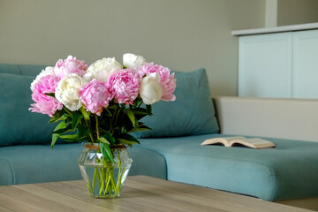 Close up shot of wooden coffee table with bouquet of beautiful white peony flowers in glass vase on foreground and blue textile couch on the background. Copy space for text, close up.