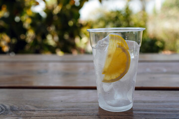 Plastic glass with ice and two slice of lemon on wooden table with blured greens on background