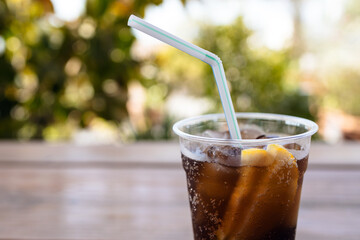 Plastic sweated glass with cold cola,  ice and two slice of lemon on wooden table with blured greens on background. Copy space.