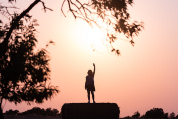 silhouette of middle-aged latina woman on top of a straw bale raising an arm with the evening sun behind her.