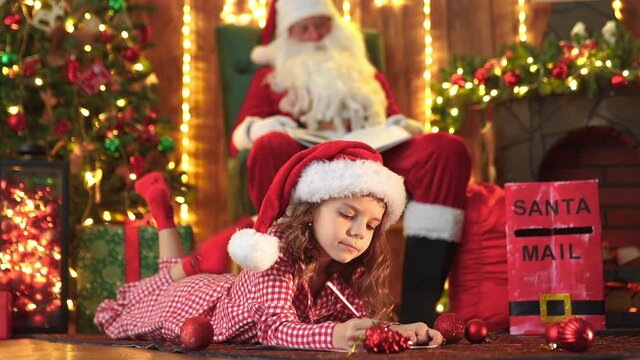 Funny girl in pajamas and Santa hat writes letter to Santa near Christmas tree.