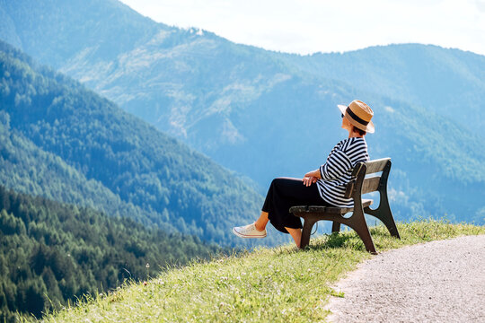 Young fashionable dressed female in straw hat sitting on a bench enjoying Santa Maddalena village view and stunning picturesque Dolomite Alps peaks in Val di Funes valley, South Tyrol, Italy.