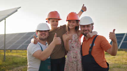 Portrait of two dads and two kids in protective helmets at a solar power plant, all pointing fingers up at the camera and smiling. Parents put a helmet on their children's heads at a solar power plant