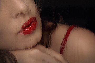 Close-up portrait of woman leeps and nose seen through wet glass. Red lips and strap of dress close...