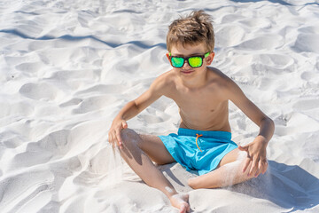 Handsome young boy in blue shorts and sunglasses sits on the white sand on the beach. Plays with white sand.