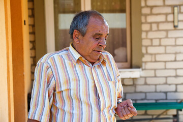 Portrait of a senior man outdoors. 70s years old latin or hispanic man standing outside in summer day and looking down on his nails. Casual and lifestyle concept