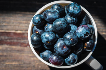 Large, juicy berries, blueberries in a light mug on a gray-cinnamon wooden surface, light shadow, sunbeam. Top view with space for text. Banner