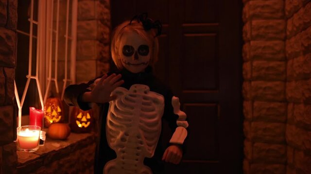 Little child in scary skeleton costume at Halloween celebration party. The child is ready for trick or treat on Halloween night. The kid waves his hand before knocking on the door of the house