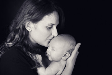 Happy woman mother kissing a newborn baby boy in her arms, black studio background. Mom with a child in her hands