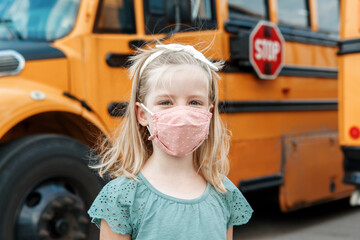 Girl kid student in protective face mask near school yellow bus outdoors. New normal at coronavirus covid-19 pandemic. Measurements against virus spread in class.