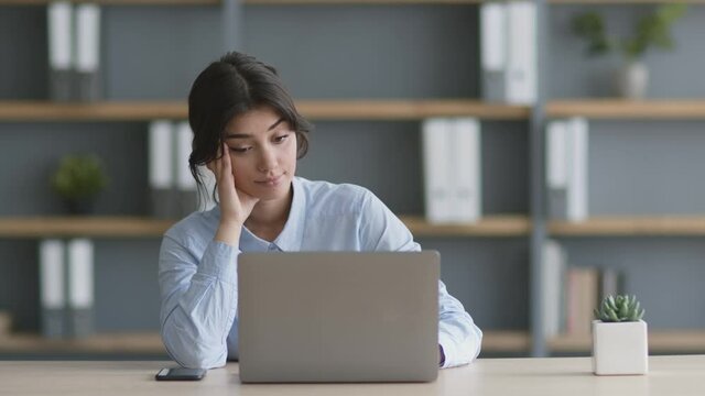 Young woman office manager sitting at workplace and web surfing on laptop, feeling boring from job, slow motion