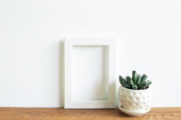 Obraz na płótnie Canvas Blank white photo frame and flowerpot on wooden shelf. white wall background. home interior