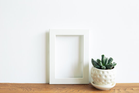 Blank white photo frame and flowerpot on wooden shelf. white wall background. home interior