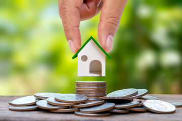 Hand-holding model house on a pile of coins with blur green natural background, home loan, and real estate investment loan.