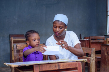 African Nigerian mother or teacher sitting together with her girl child in a classroom, helping her...